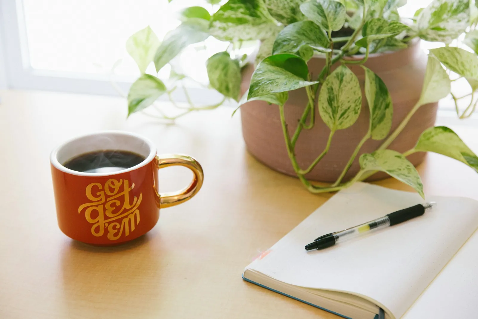 Kaffeetasse, aufgeschlagenes Notizbuch und eine Pflanze auf einem hellen Schreibtisch